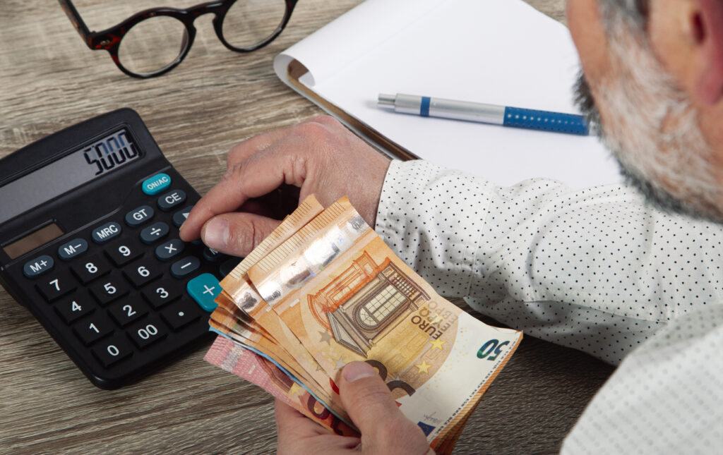 Imagem de duas mãos masculina segurando um montante de dinheiro e utilizando uma calculadora que está sobre uma mesa de madeira com vários papéis